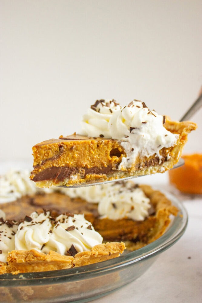 A slice of pumpkin pie with a chocolate layer is being lifted from a pie dish, topped with whipped cream and chocolate shavings.