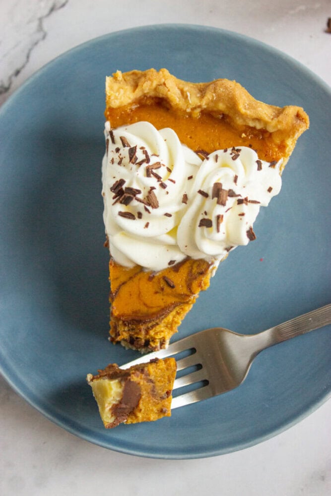 A slice of pumpkin pie topped with whipped cream and chocolate shavings on a blue plate, with a fork holding a bite-sized piece.