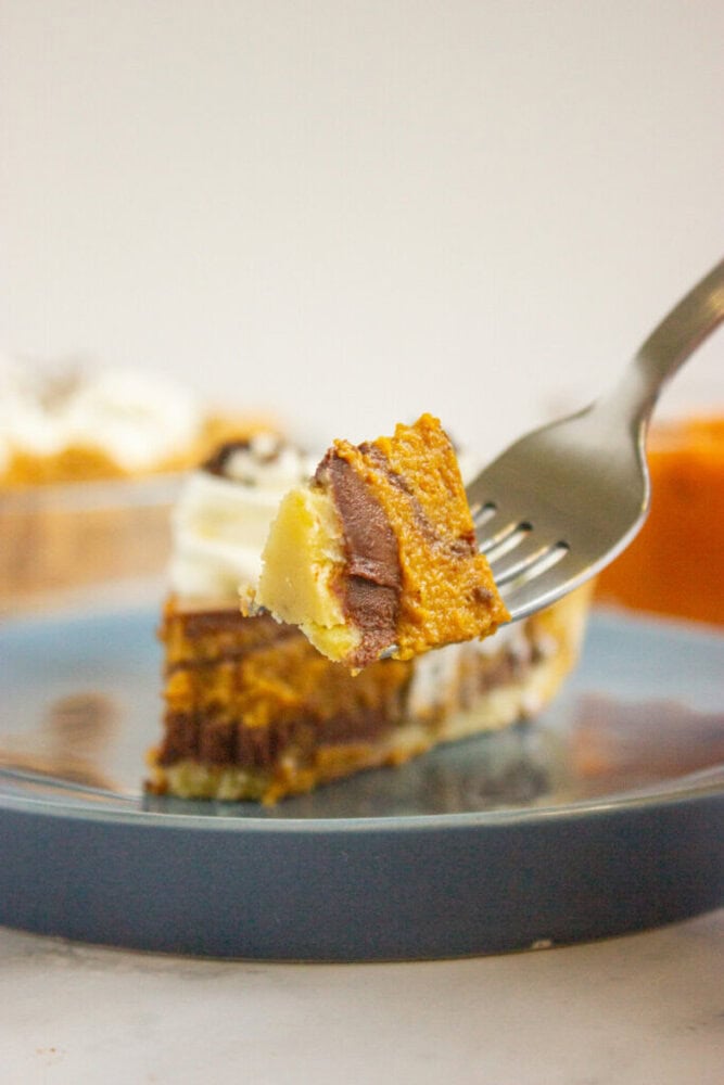 A fork holds a bite of layered chocolate and pumpkin cheesecake above a blue plate with the remaining slice in the background.