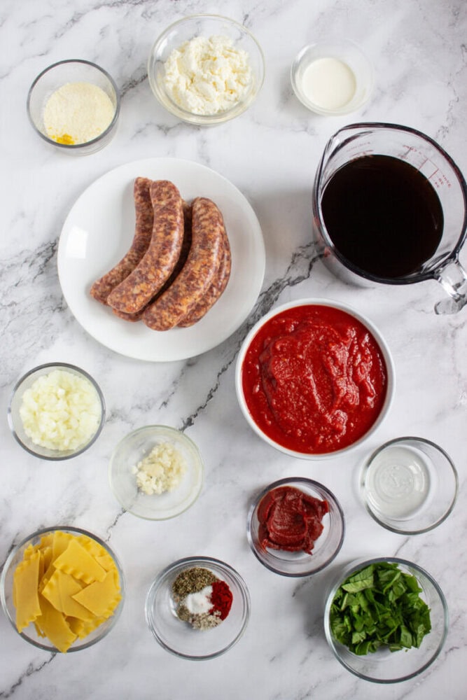 A variety of ingredients in small bowls on a marble surface, including uncooked sausages, tomato sauce, cheese, onions, garlic, herbs, red wine, and seasonings.