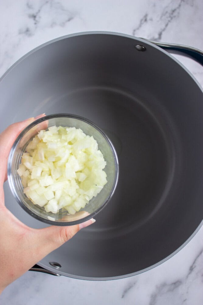 A hand holds a bowl of chopped onions over an empty gray pot on a marble countertop.