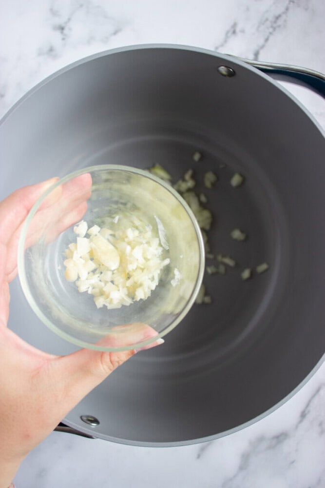A hand pours minced garlic from a small glass bowl into a large empty pot on a marble countertop.