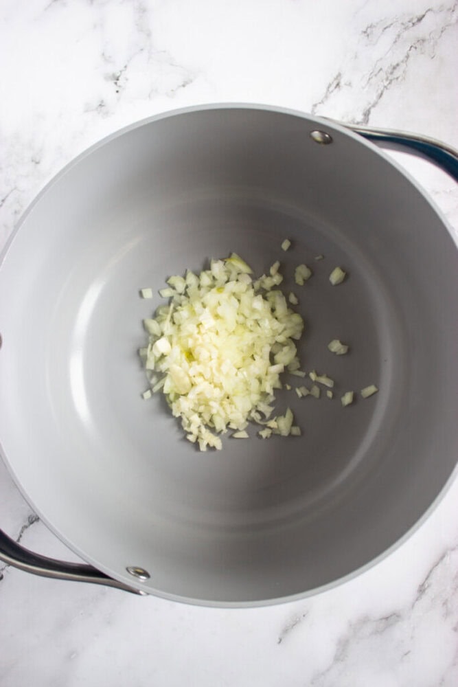 Diced onions in the center of a large, empty, gray cooking pot on a white marble surface.