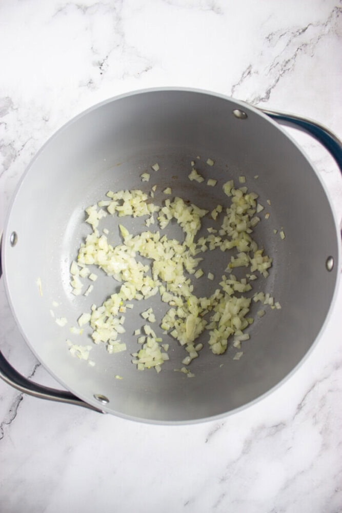 Chopped onions and minced garlic sautéing in a large, empty pot on a white marble countertop.