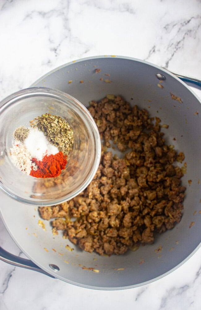 A glass bowl with assorted spices is held above a pot of cooked ground meat on a marble countertop.