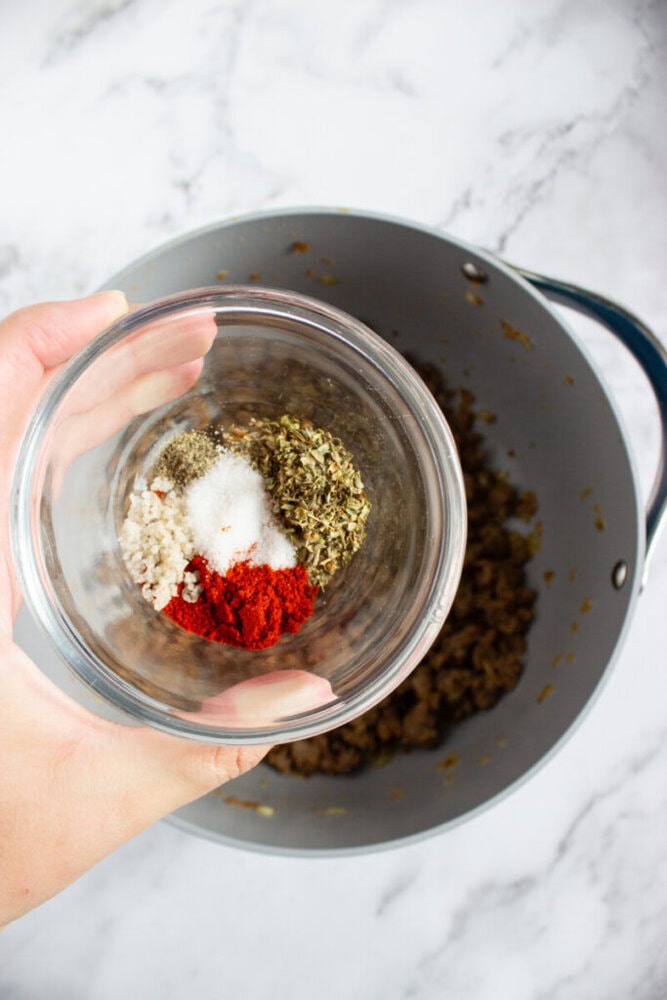 A hand holding a glass bowl containing paprika, oregano, salt, black pepper, and coarse salt over a pot with cooked ingredients on a marble surface.