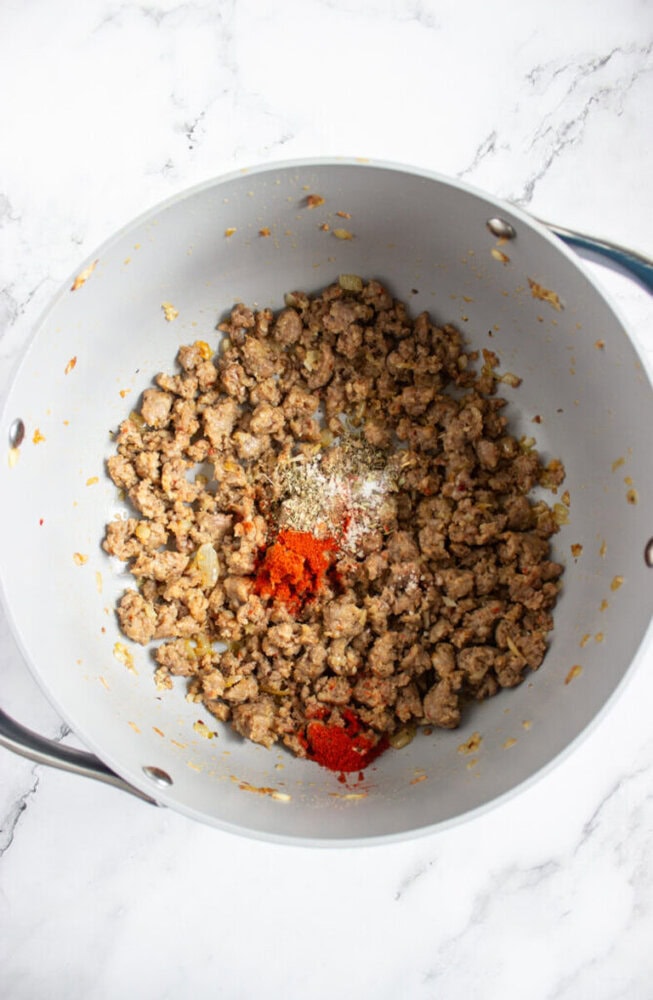 Ground meat is being cooked in a pot with visible dried herbs and red spices added on top, on a white marble surface.