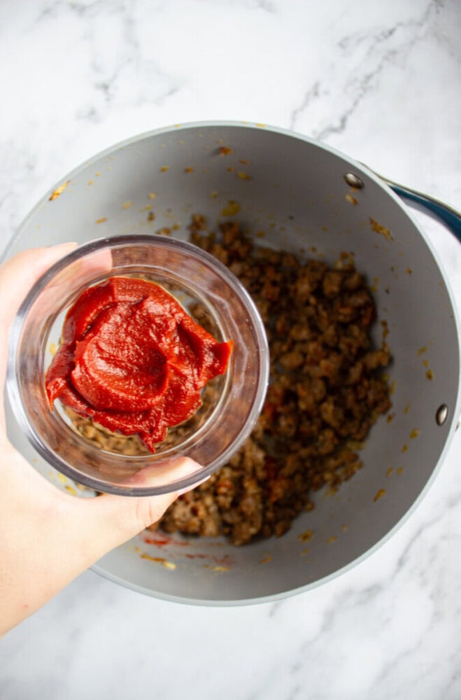 A hand holds a small glass bowl of tomato paste above a pot containing browned ground meat on a marble countertop.