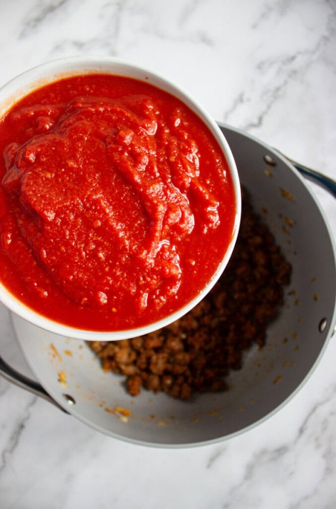 A bowl of tomato sauce is being poured into a pot containing cooked ground meat.