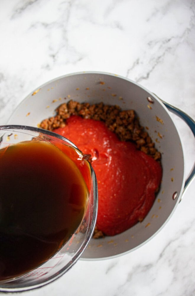 A glass measuring cup pours brown liquid into a pot containing ground meat and tomato sauce, on a white marble surface.