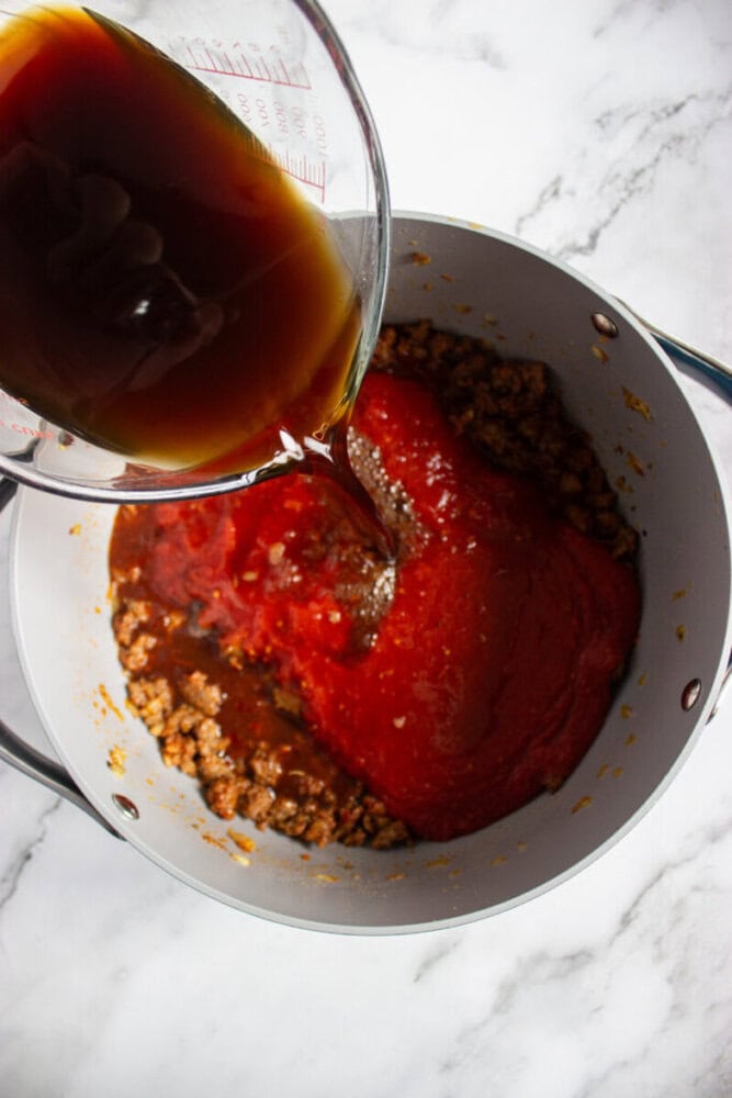 A measuring cup pours dark liquid into a pot containing ground meat and tomato sauce on a white marble countertop.