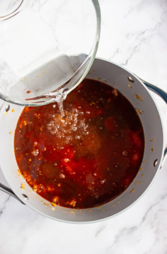 A glass container pours liquid into a pot filled with a red, tomato-based mixture on a marble countertop.