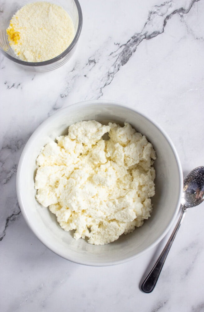 A white bowl filled with ricotta cheese next to a spoon on a marble surface, with a glass bowl of grated cheese in the background.