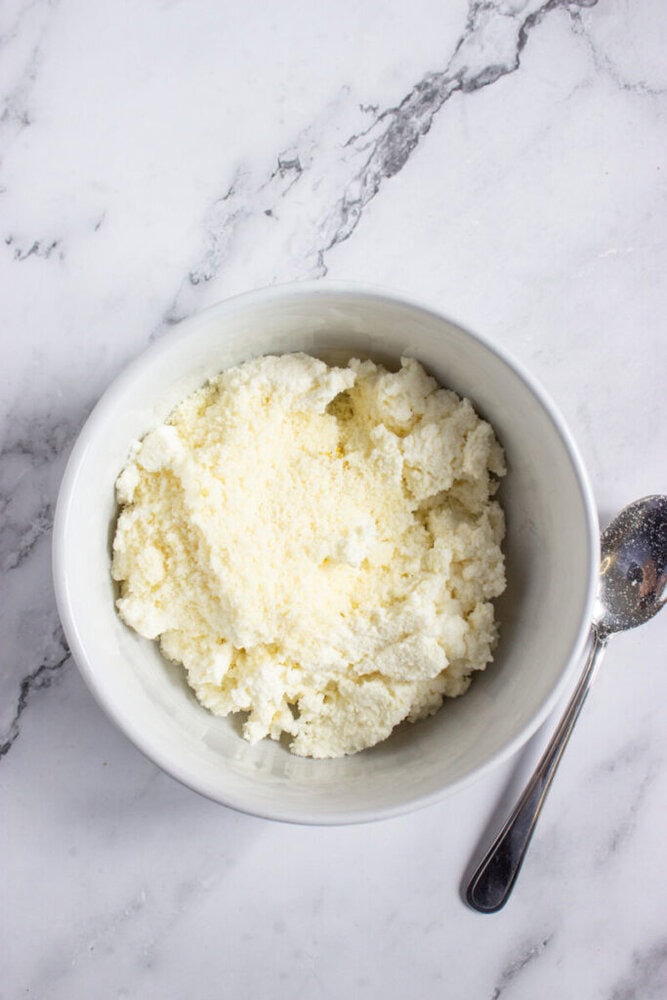 A white bowl filled with ricotta cheese sits on a marble surface next to a metal spoon.