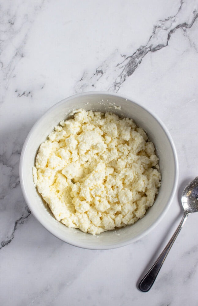 A white bowl filled with crumbled white cheese sits on a marble surface next to a metal spoon.