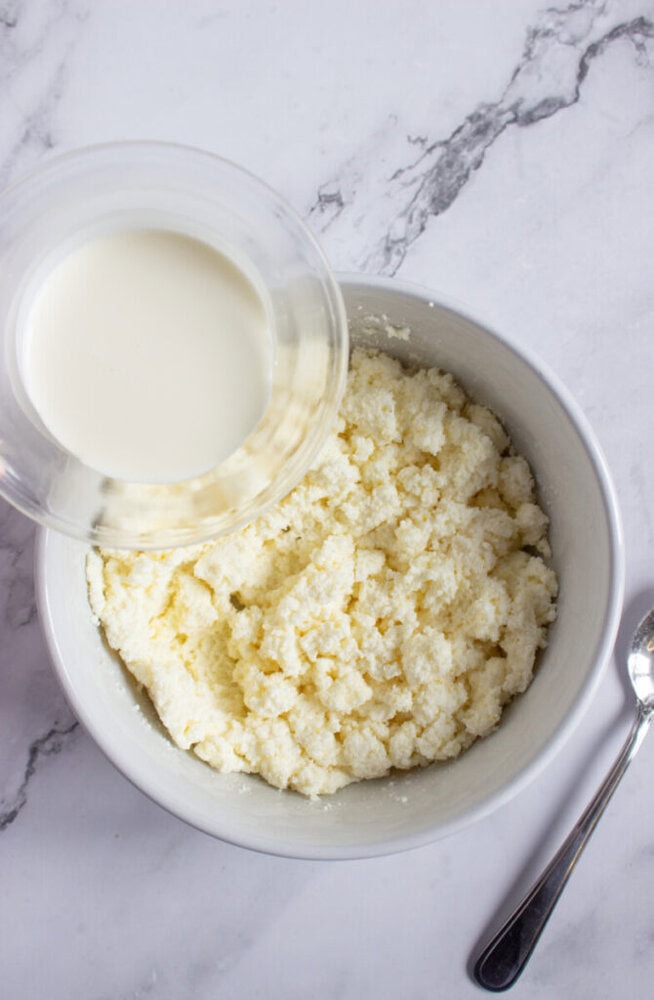 A bowl of crumbled cheese next to a spoon, with a glass container of milk being poured in on a marble surface.