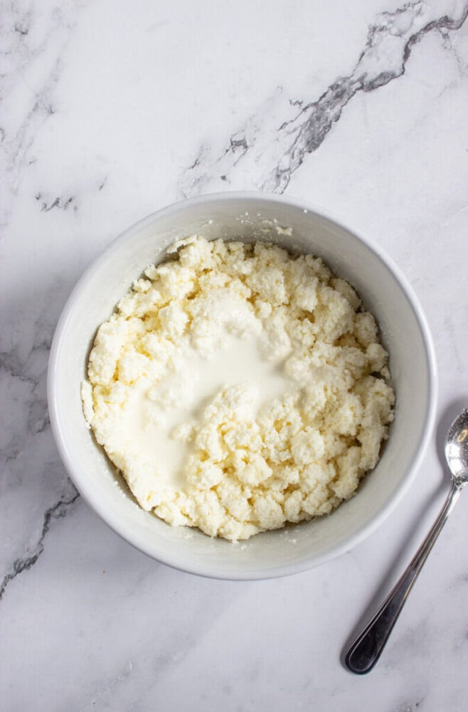 A bowl containing crumbled cheese with cream on top, placed on a white marble surface next to a metal spoon.