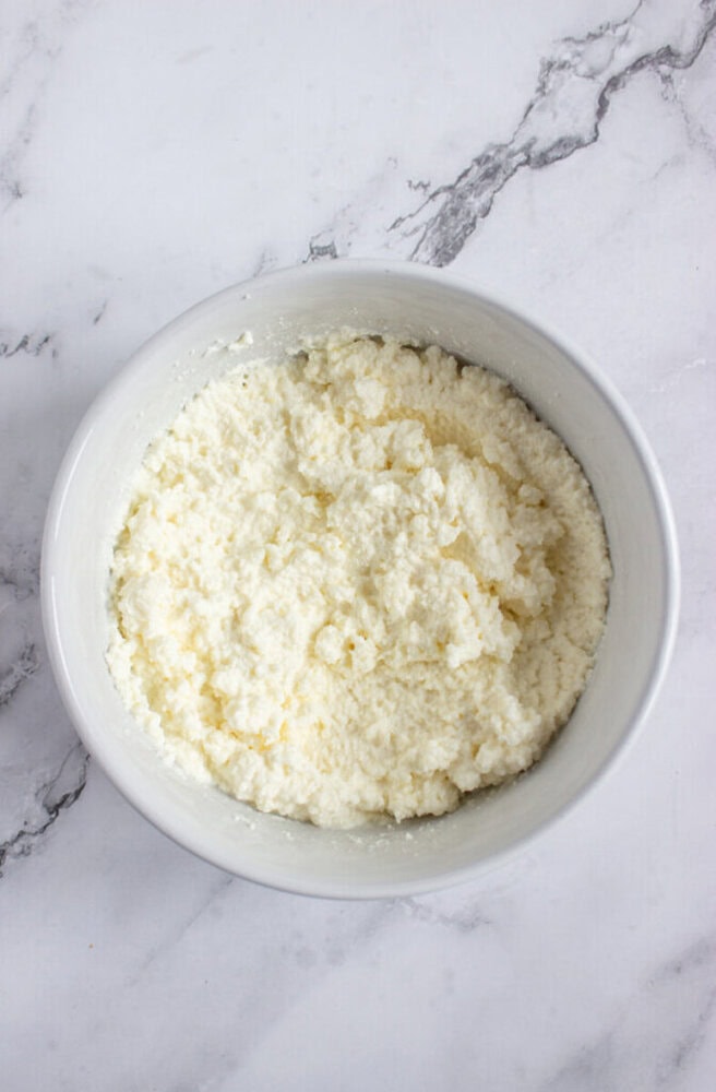 A white bowl filled with crumbled cottage cheese sits on a white marble surface.