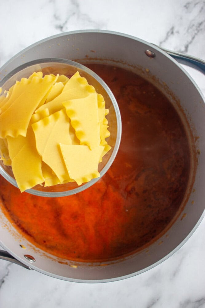A bowl of uncooked lasagna noodles is held over a pot of red tomato sauce on a marble countertop.