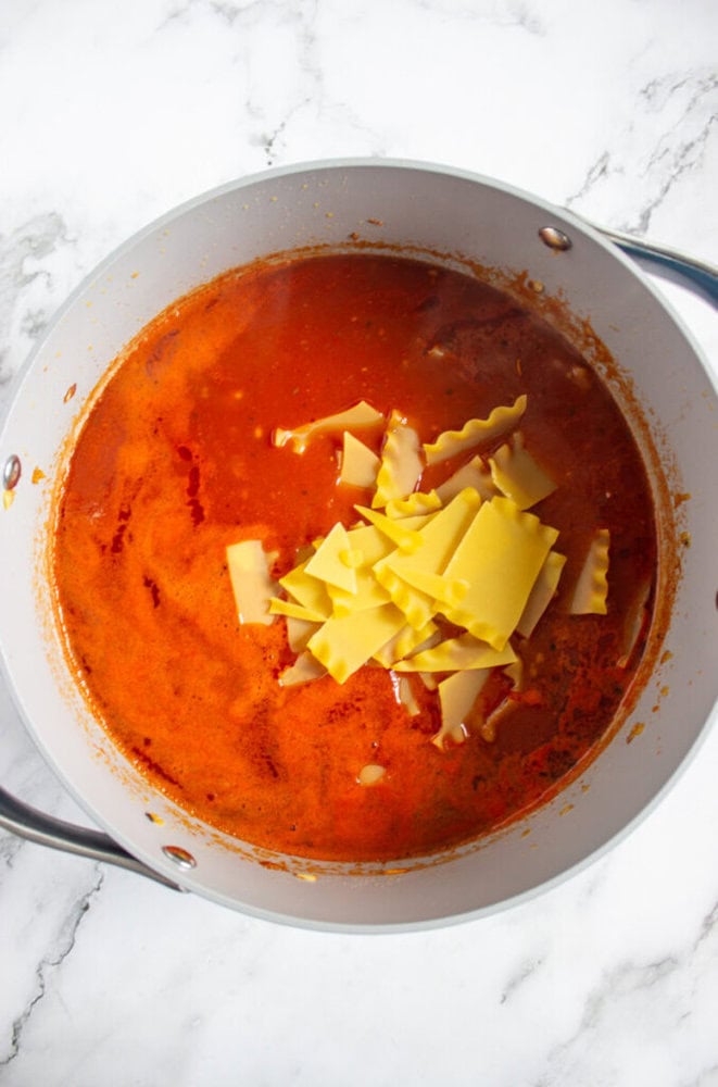 A pot of tomato-based soup or sauce with broken pieces of uncooked lasagna noodles on top, placed on a white marble surface.