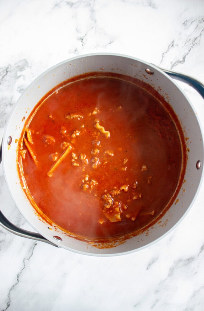 A pot of red tomato-based sauce with ground meat and broken lasagna noodles simmering, viewed from above on a marble surface.