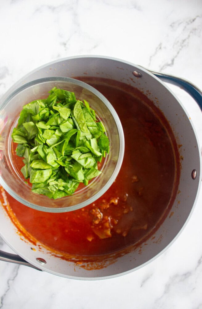 A bowl of chopped spinach is being held above a pot of tomato-based sauce with visible meat chunks on a marble countertop.
