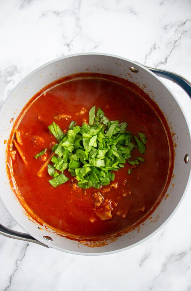 A pot of red tomato-based soup or stew with chopped green leafy vegetables added on top, on a white marble surface.