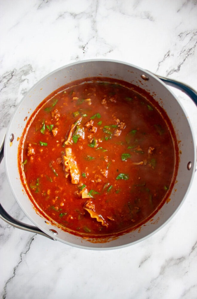 A pot filled with red tomato-based soup or stew containing ground meat, broken pasta, and chopped greens, placed on a white marble surface.