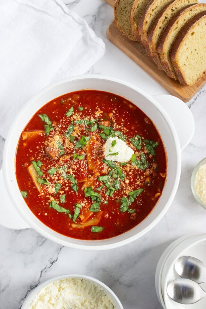 A white bowl of tomato-based soup topped with cheese and herbs sits on a marble surface, with sliced bread and bowls of grated cheese nearby.