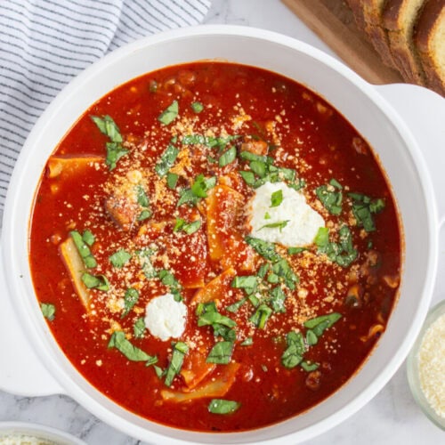 A bowl of red tomato-based soup topped with cheese and herbs sits on a marble surface, surrounded by sliced bread and small bowls of grated cheese.