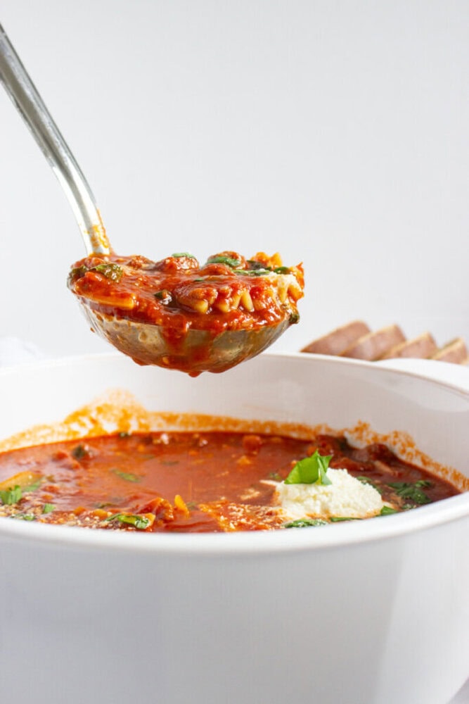 A ladle lifts a serving of tomato-based soup with pasta and greens from a white bowl; a dollop of ricotta and basil garnish are visible.