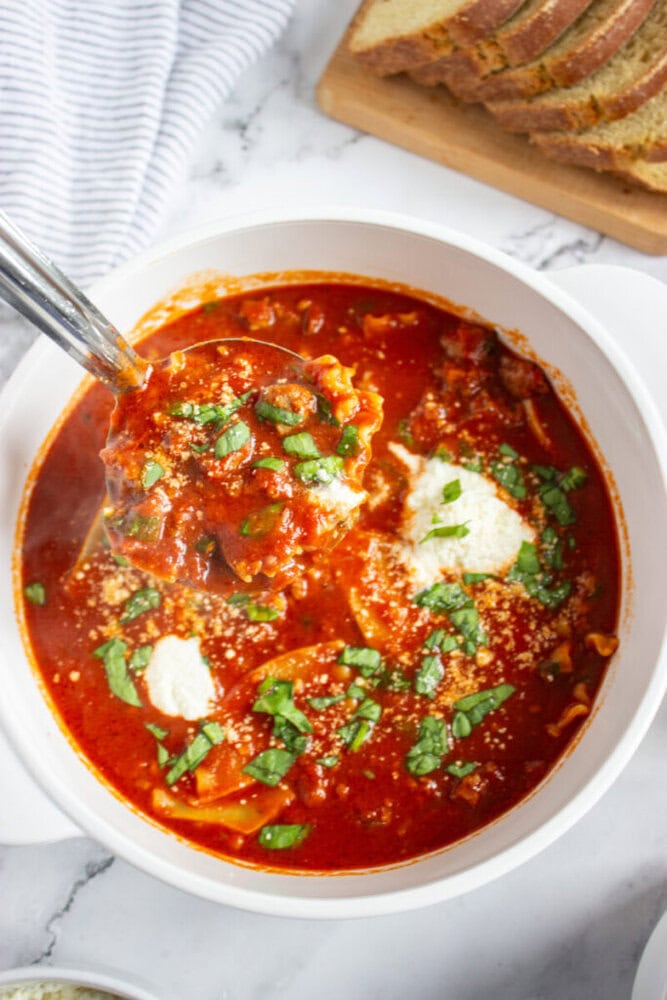 A bowl of tomato-based soup with lasagna noodles, ricotta cheese, and fresh basil, being served with a ladle. Sliced bread is visible in the background.