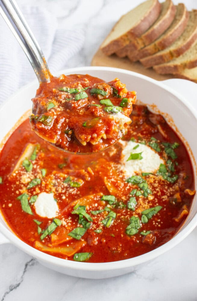 A ladle is lifting lasagna soup from a bowl, with visible noodles, tomato sauce, cheese, and fresh basil. Slices of bread are on a cutting board in the background.