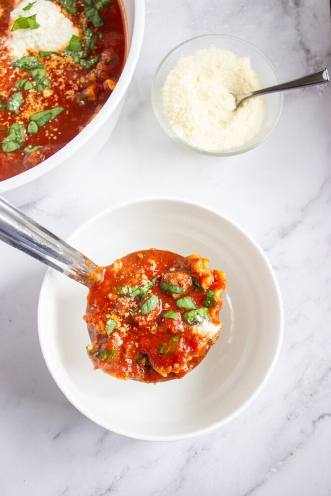 A ladle of tomato-based soup with spinach and grated cheese is being served into a white bowl, with a bowl of grated cheese and more soup in the background.
