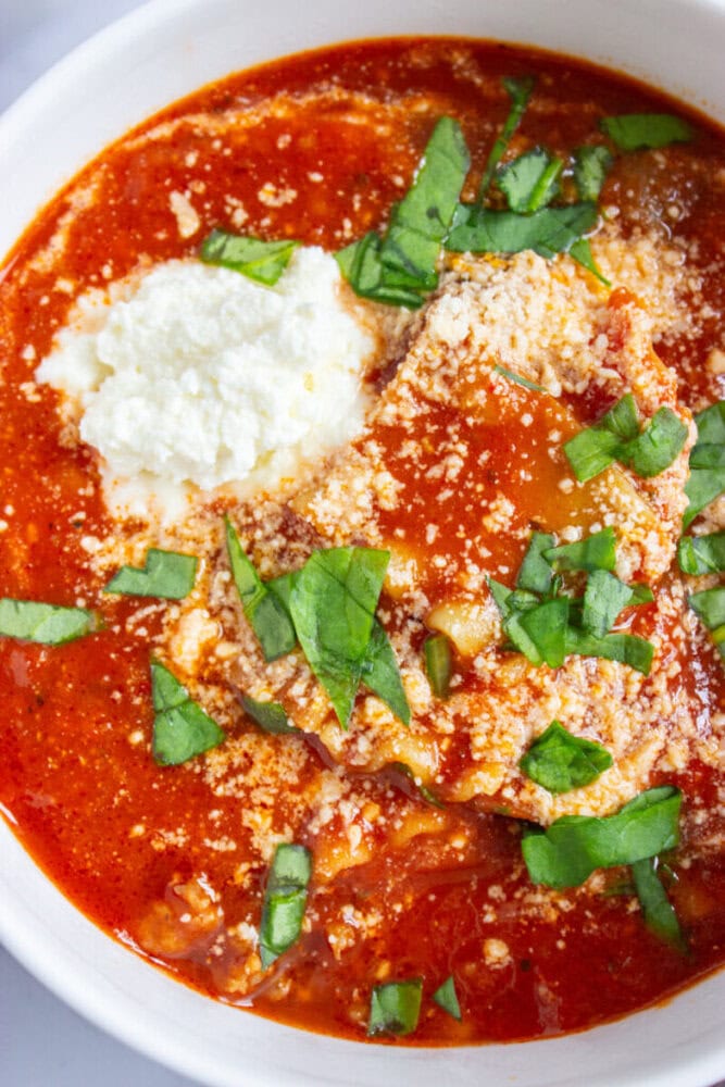 A bowl of lasagna soup topped with ricotta cheese, grated Parmesan, and chopped fresh basil.