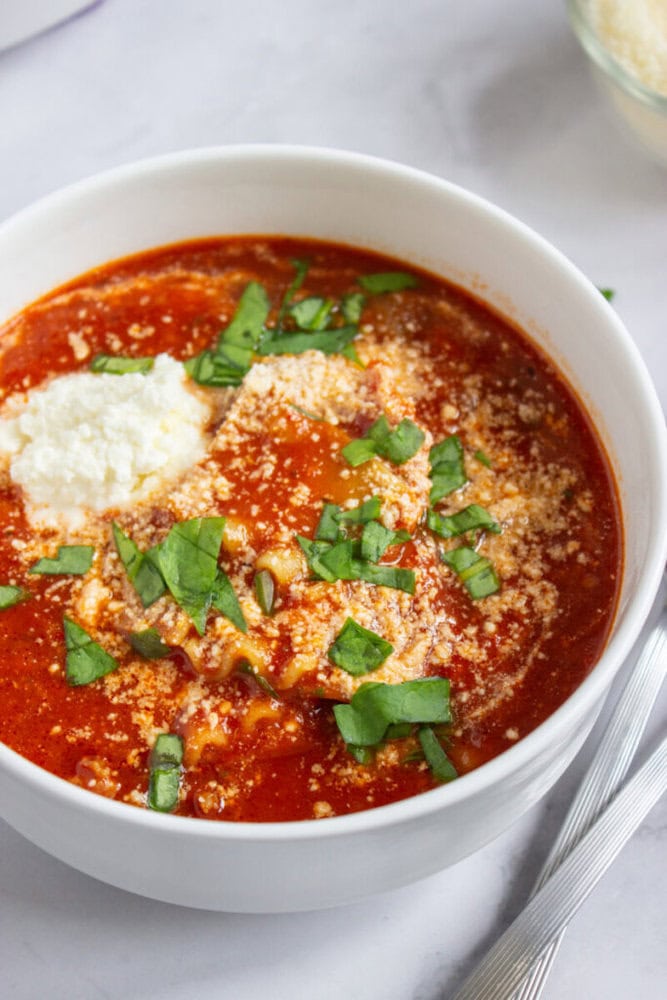 A white bowl filled with red soup topped with ricotta cheese, grated Parmesan, and chopped fresh basil, placed on a white surface with a spoon beside it.