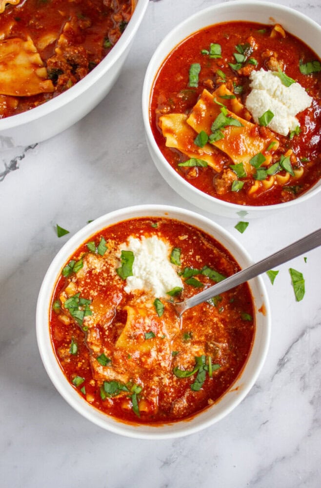 Three white bowls filled with lasagna soup, topped with a dollop of ricotta cheese and chopped fresh herbs, are placed on a marble surface.