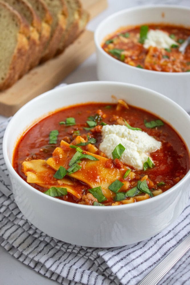 A bowl of lasagna soup topped with ricotta and chopped basil, placed on a striped cloth with a second bowl and sliced bread in the background.
