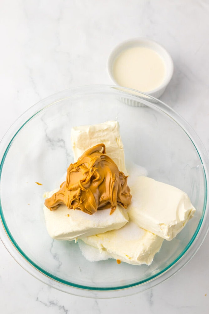 A glass bowl with cream cheese blocks, sugar, and peanut butter, next to a small dish of cream on a marble surface.