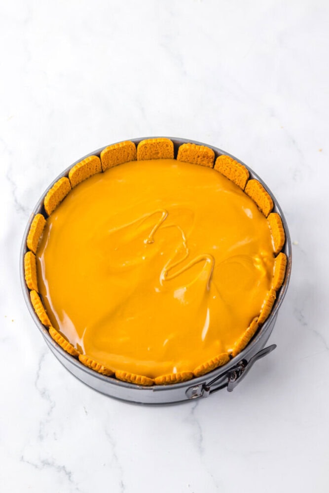 A round cheesecake with a smooth orange topping and a crust made of rectangular cookies arranged vertically around the edge, in a springform pan on a white surface.