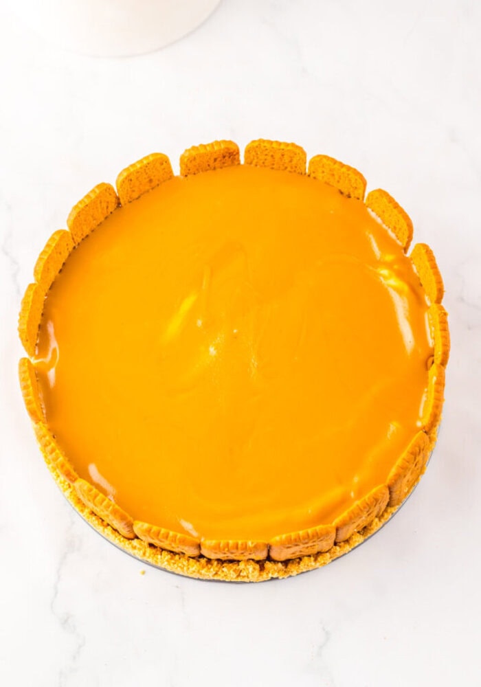 A round cheesecake with a glossy orange topping and cookies arranged vertically around the edge, on a white surface.