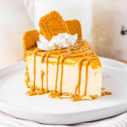 A slice of cheesecake with caramel drizzle, whipped cream, and two rectangular cookies on top, served on a white plate.