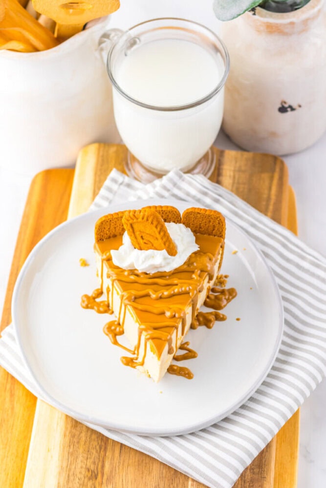 A slice of caramel-drizzled cheesecake topped with whipped cream and cookies on a white plate, next to a glass of milk on a wooden board.
