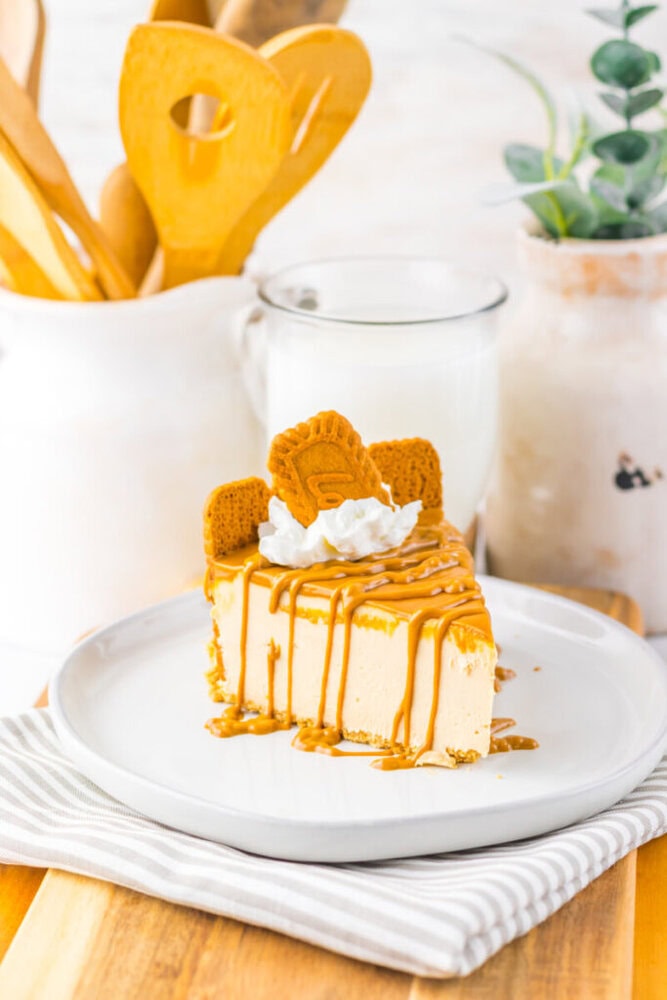 A slice of cheesecake topped with caramel drizzle, whipped cream, and two cookies, served on a white plate with a glass of milk in the background.