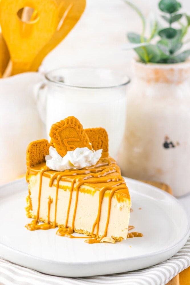A slice of cheesecake topped with caramel drizzle, whipped cream, and two cookies, served on a white plate with a glass of milk and decorative items in the background.