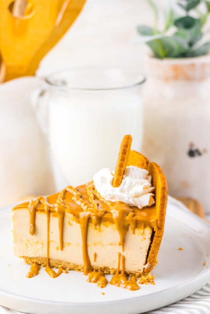 A slice of cheesecake with caramel sauce, whipped cream, and a cookie, served on a white plate with a glass of milk in the background.
