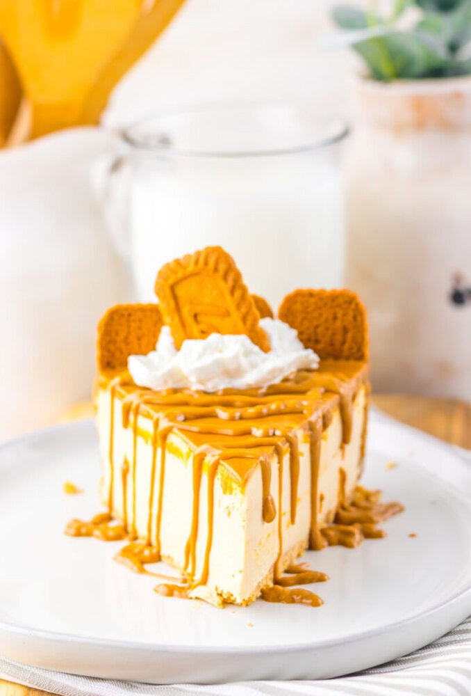 A slice of cheesecake topped with caramel sauce, whipped cream, and three rectangular cookies, served on a white plate with a glass of milk in the background.