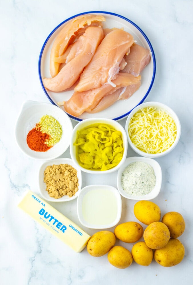 Raw chicken breasts on a plate with bowls of shredded cheese, ranch seasoning, sliced pepperoncini, spices, milk, brown sugar, butter, and small yellow potatoes on a white surface.