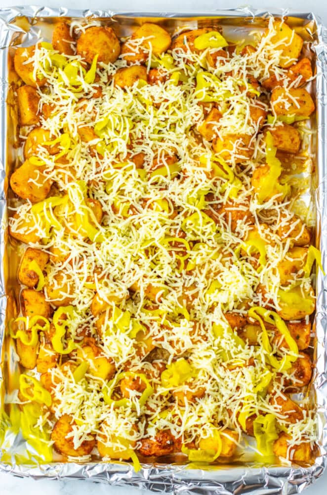 Sliced potatoes topped with shredded cheese and banana pepper rings spread on a foil-lined baking sheet.