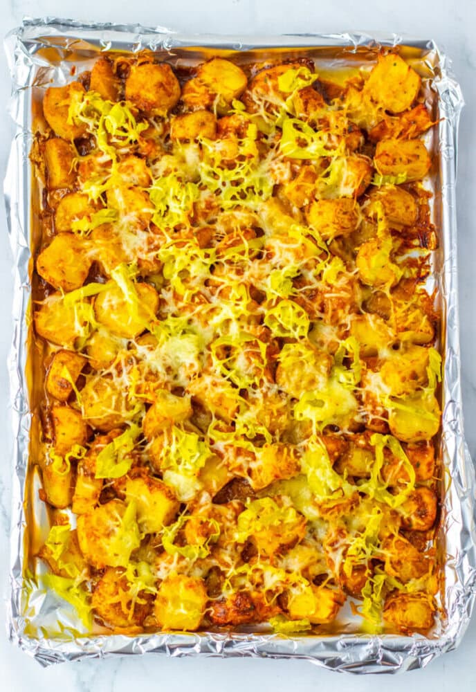 Sheet pan with roasted potato slices, shredded cheese, and sliced yellow peppers on aluminum foil, photographed from above.